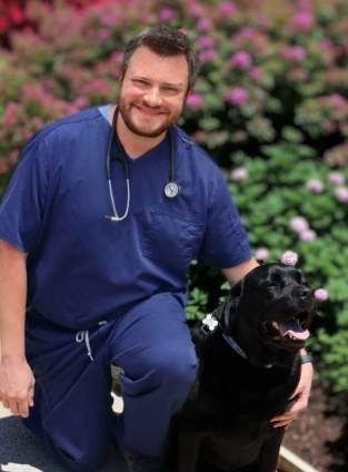 Dr. Peter Beron smiling in blue scrubs kneeling beside a black Labrador among flowering bushes