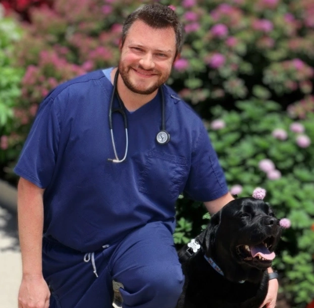 Dr. Peter Beron smiling in blue scrubs kneeling beside a black Labrador among flowering bushes