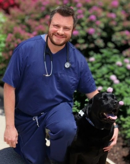 Dr. Peter Beron smiling in blue scrubs kneeling beside a black Labrador among flowering bushes