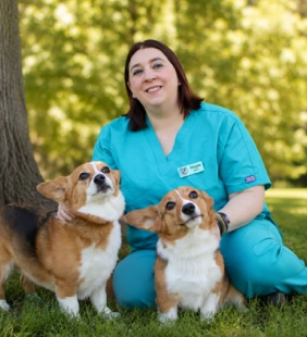 Staff member in teal scrubs kneeling in grass with two corgis