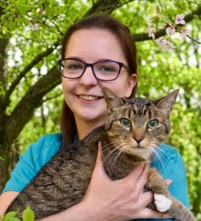 Staff member in teal scrubs smiling and holding a tabby cat outdoors
