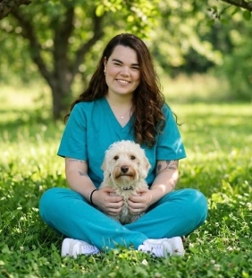 Staff member in teal scrubs sitting in grass with a curly-haired white dog